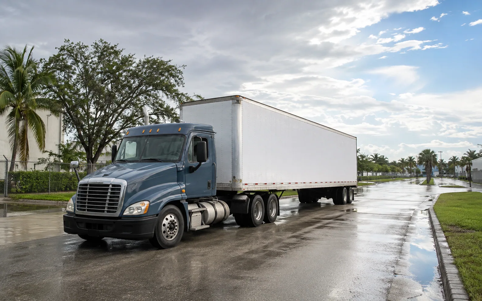 Illustrative view of an unbranded tractor-trailer on a wet palm-lined industrial road after rain.